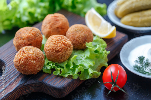 Falafel lies on a wooden cutting board. On the table lie tomatoes, cucumbers, lettuce, dill, lemon, sour cream.