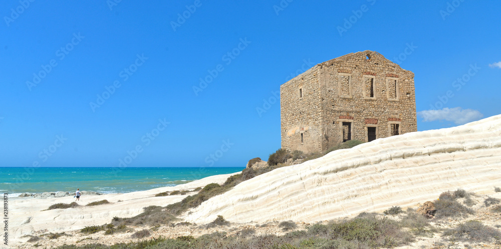 Woman on empty white beach with old ruins of abandoned stone house on the rocks and summer blue sky and sea in background in Sicily Italy