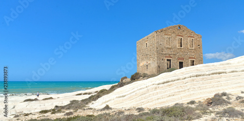 Woman on empty white beach with old ruins of abandoned stone house on the rocks and summer blue sky and sea in background in Sicily Italy