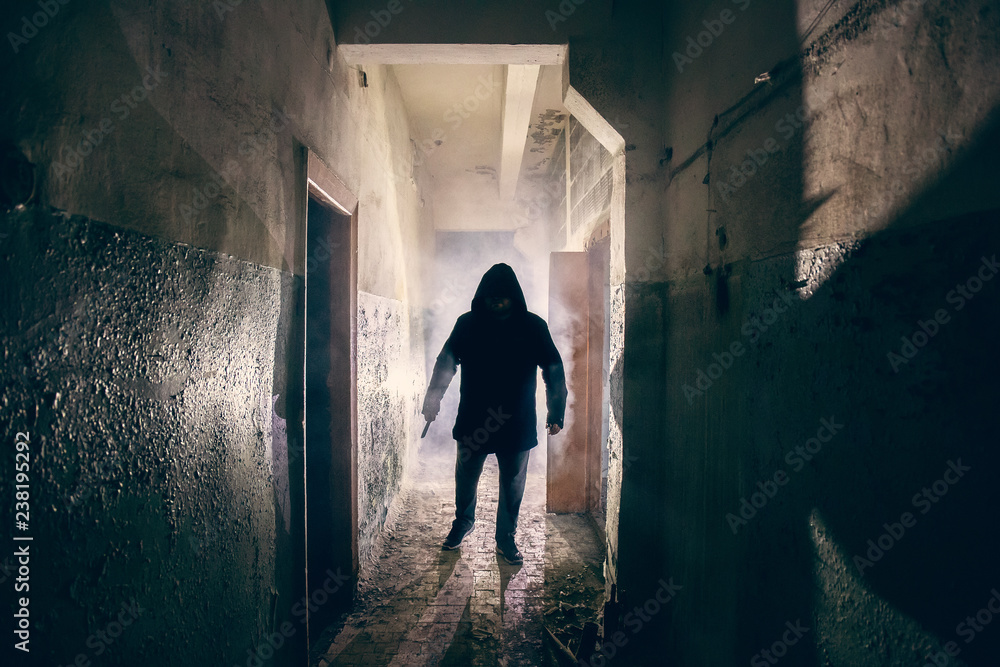 Dark silhouette of strange danger man with knife in hand in hood in back light with smoke or fog in scary grunge corridor or tunnel