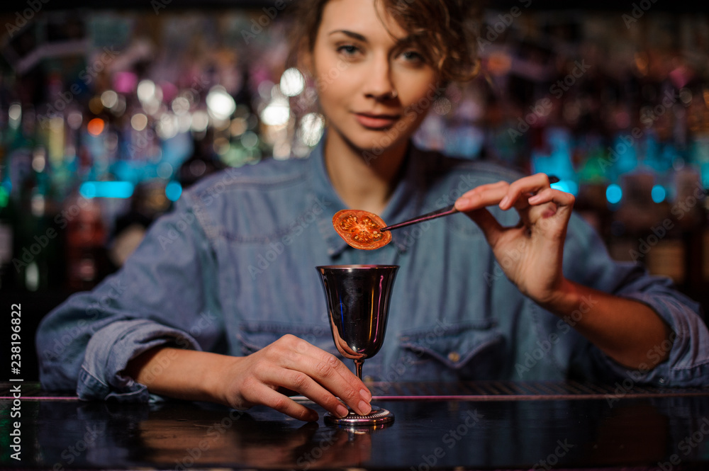 Smiling female bartender adding a dried tomato with tweezers to a cocktail glass with alcoholic drink
