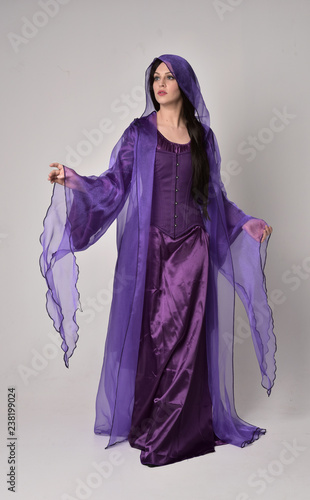 full length portrait of beautiful girl with long black hair, wearing purple fantasy medieval gown and cloak. standing pose on grey studio background.