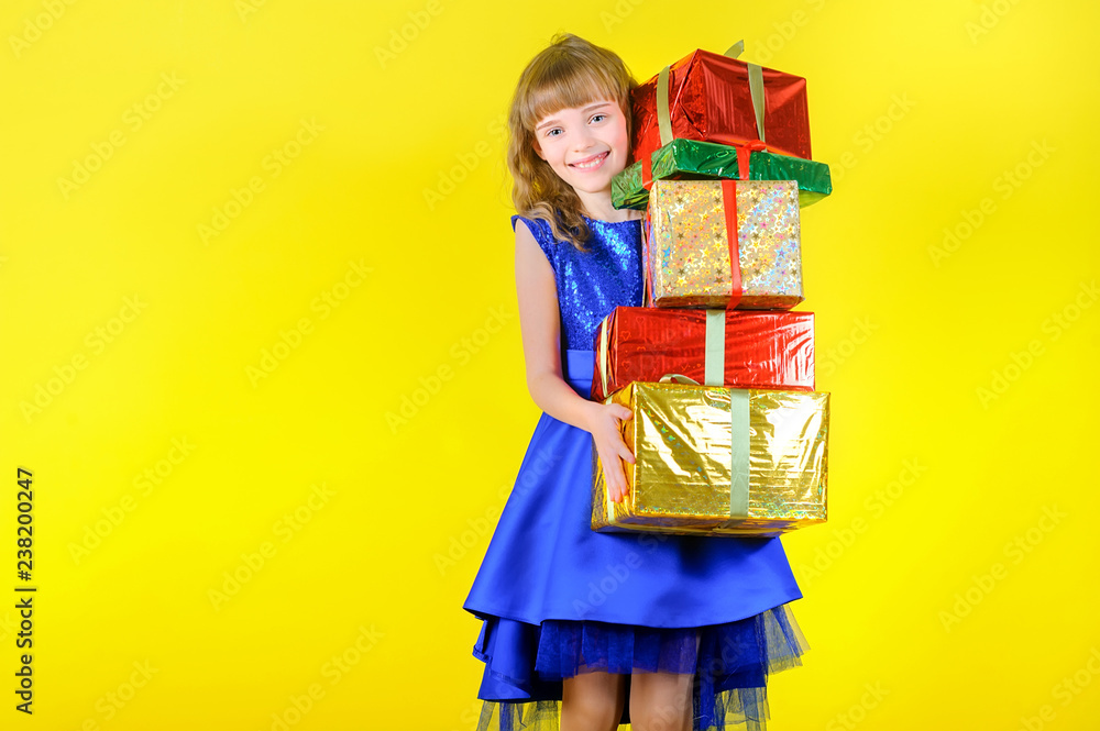 festive emotions, beautiful young girl in a festive blue dress holding a lot of gifts and smiling, in the studio on a yellow background