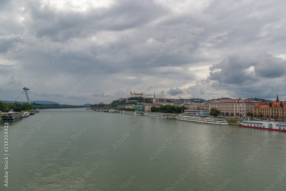 Panoramic view in a cloudy day of the Danube River with moored cruise ships, the Ufo(Most SNP) Bridge , Cathedral of Saint Martin and the Bratislava Castle in background.
