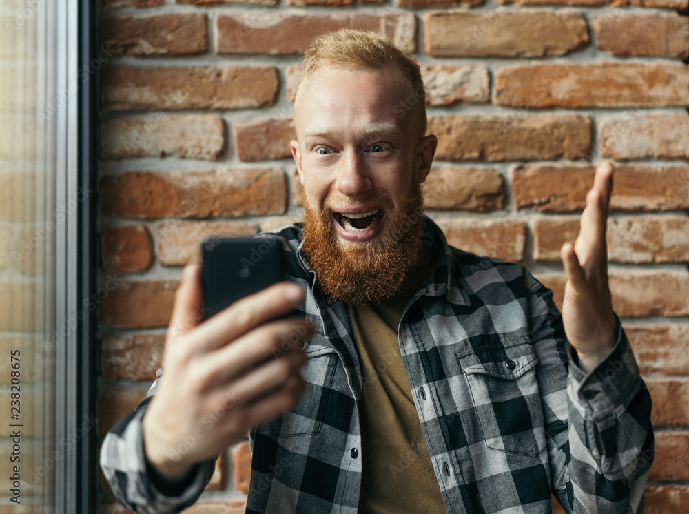 Overjoyed man hipster with happy emotion face wide open mouth and eyes using mobile application for online shopping with discount sales low price. Cash back. Bearded guy celebration victory - obrazy, fototapety, plakaty 