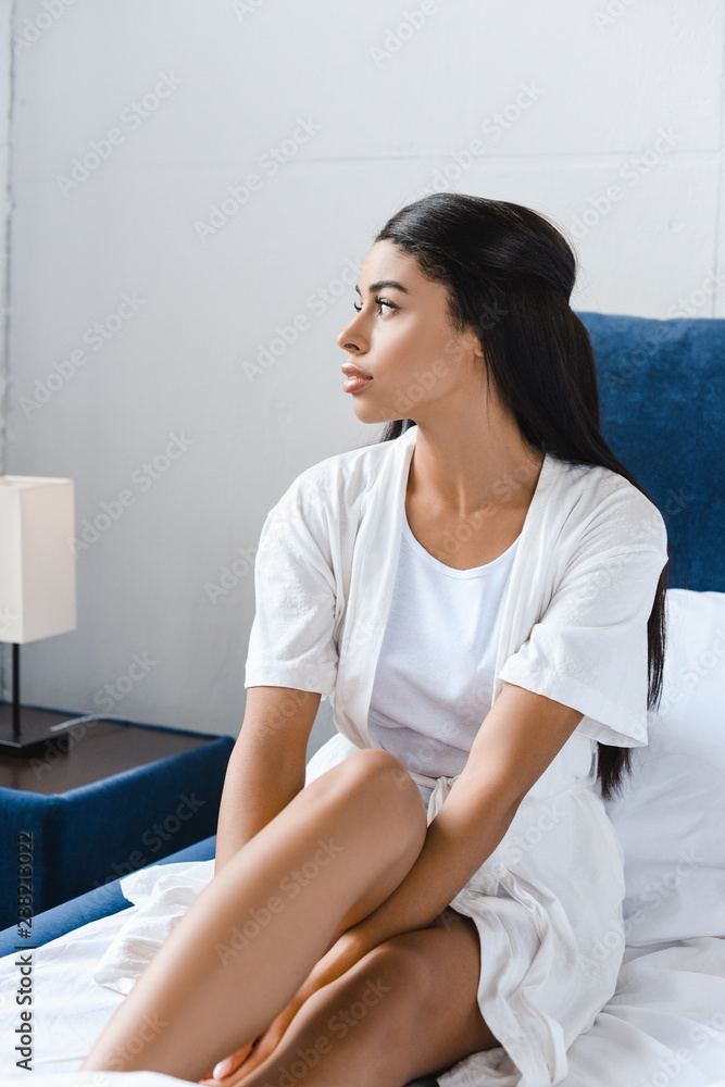 attractive mixed race girl in white robe sitting on bed and looking away attractive mixed race girl in white robe sitting on bed and looking away