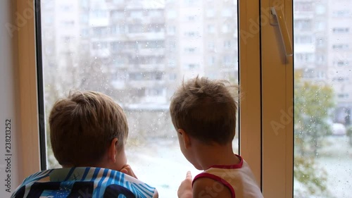 Children stand at the window and watch the snow falling on the street. First day of winter