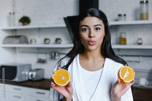 beautiful mixed race girl in white robe holding cut orange, grimacing and looking at camera in morning in kitchen