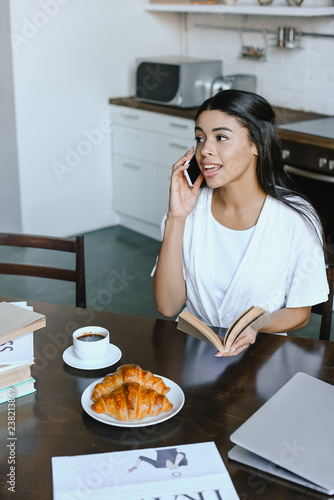 smiling attractive mixed race girl in white robe talking by smartphone in morning in kitchen and holding book