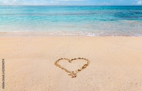 A heart drawn in the white sand of a Caribbean beach  with turquoise blue ocean in the background.