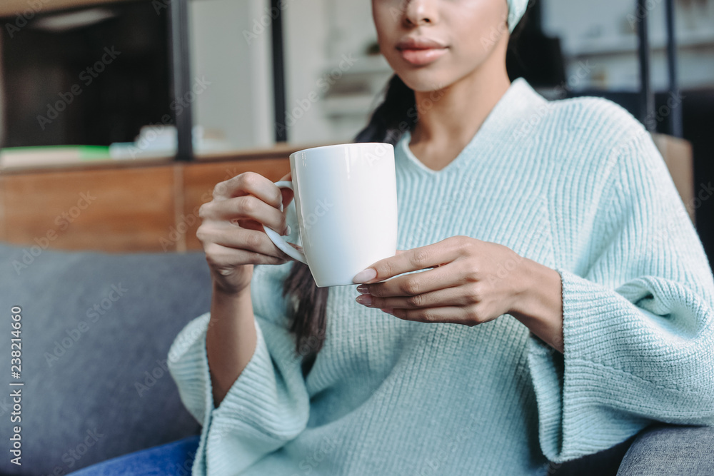 cropped image of mixed race girl in turquoise sweater and headband sitting on sofa and holding cup of tea in living room cropped image of mixed race girl in turquoise sweater and headband sitting on sofa and holding cup of tea in living room