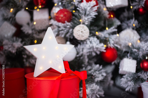 White glowing LED star on red gift box near decorated Christmas tree.