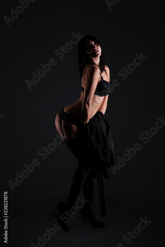 Sexy young brunette woman in black sensual lingerie posing on black background wall in studio.