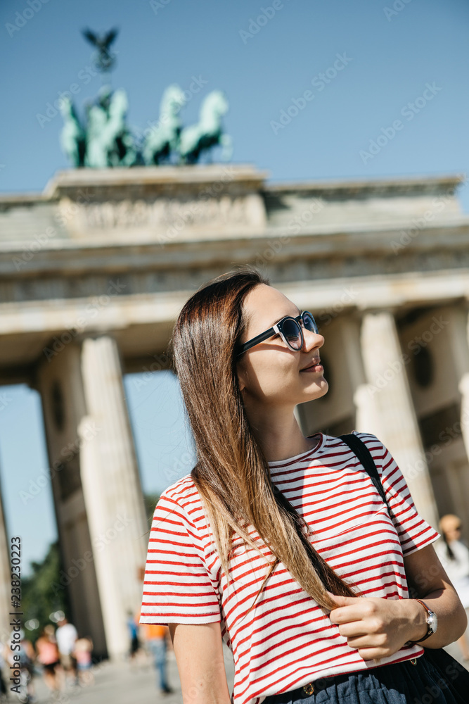 Portrait of a young beautiful positive smiling stylish tourist girl in Berlin in Germany. Brandenburg Gate and unrecognizable blurred people in the background.