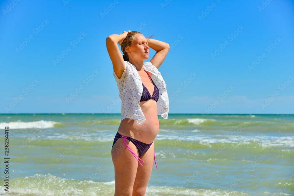A happy and joyful pregnant woman stands by the sea and ocean on the beach under the sun in the open air, hair develops in the wind. Hands holding up and enjoying the sunshine. A happy and joyful pregnant woman stands by the sea and ocean on the beach under the sun in the open air, hair develops in the wind. Hands holding up and enjoying the sunshine.
