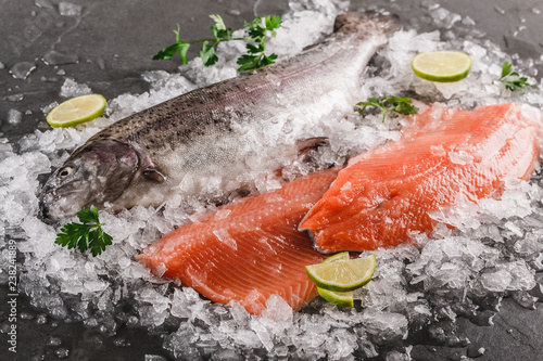 Fresh raw trout fish steak and whole fish with spices on ice over dark stone background, closeup