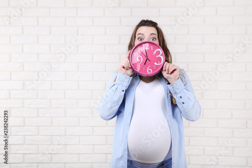 Beautiful pregnant woman holding pink clock on brick wall background