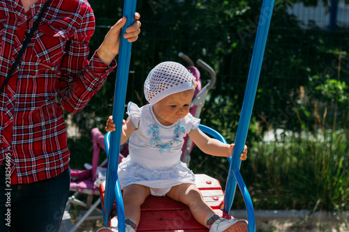 a little girl sits in the arms of a girl, she is hugged behind her back and holding hands.little girl in a white dress and hat riding on a swing, summer sun and heat. playground. childhood serenity