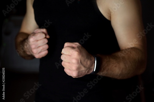 Male hands in a boxing rack with fists in a black T-shirt on a dark black background, as a symbol of the fight 