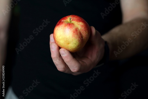 Male hand holds and gives a fresh juicy red apple, offering healthy nutrition food and diet, on a black background