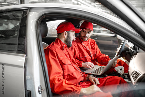 Two male auto mechanics in red uniform diagnosing car with computer sitting on the seats indoors at the car service