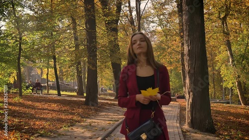 Thoughtful attrractive female walking in the park with a yellow leaf in her hand and looking around in a sunny autumn day photo