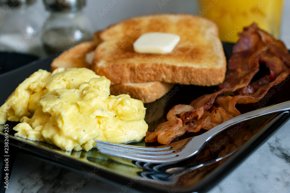 Scrambled eggs with bacon and toast with fork on black plate. Salt, pepper, and orange juice in the background. Scrambled eggs with bacon and toast with fork on black plate. Salt, pepper, and orange juice in the background.