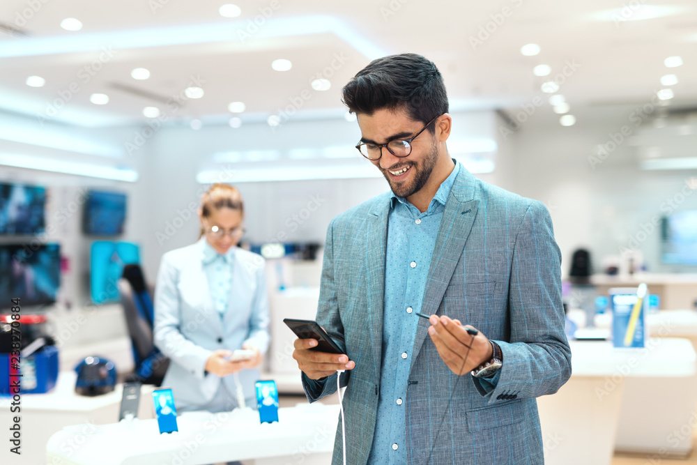 Young mixed race man dressed smart casual and with eyeglasses trying out smart phone. Tech store interior. Young mixed race man dressed smart casual and with eyeglasses trying out smart phone. Tech store interior.