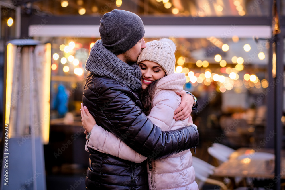A young romantic couple wearing warm clothes hugging together in evening street near a cafe outside at Christmas time