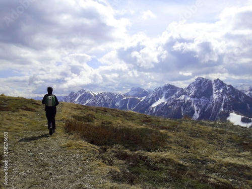 persona che cammina su sentiero di alta montagna delle alpi con sfondo dolomiti innevate e nuvole in cielo