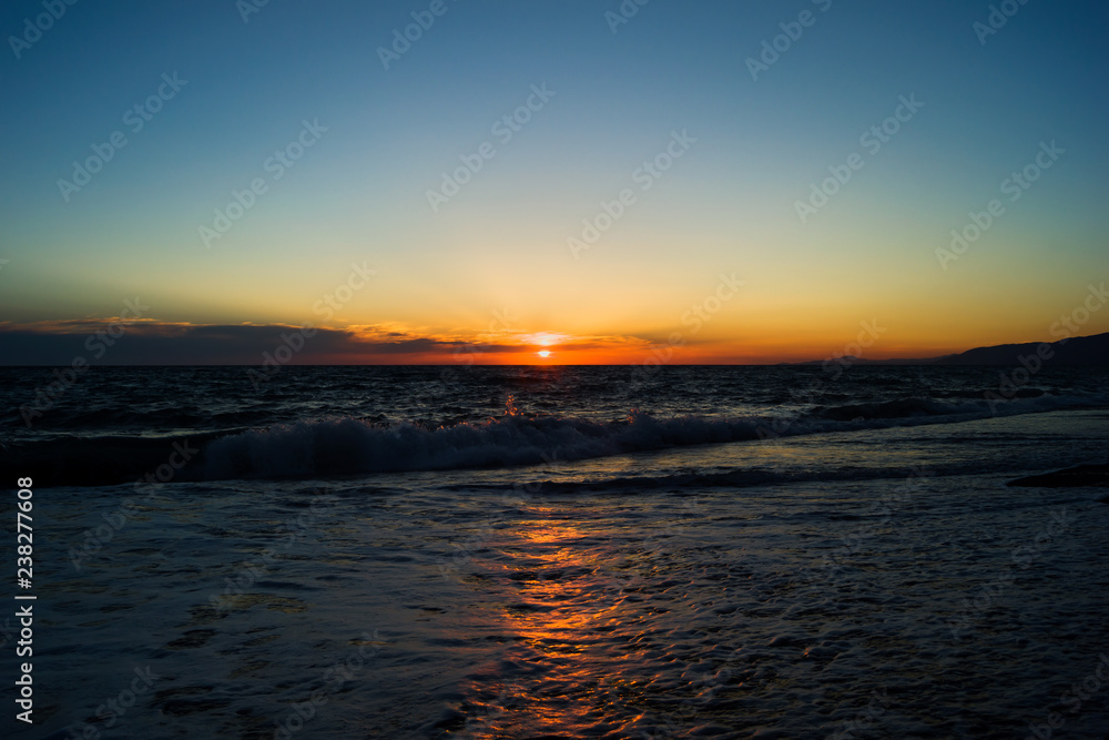Evening sun during sunset at sea. Beach with small waves and foam. Reflection of sunlight in the water.
