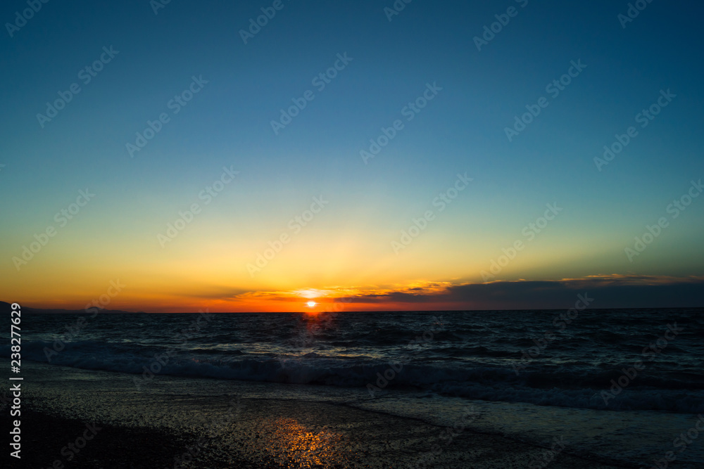 Bright and beautiful sunset on the sea. The sun's rays make their way through the clouds. Small clouds on the horizon. Pure blue sky. Reflection of sunlight in the water.