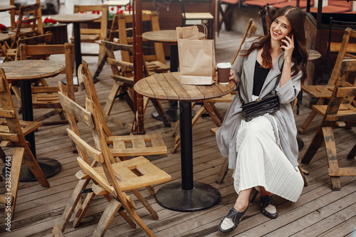 Gorgeous young woman talking on phone and smiling  with cup of coffee and paper bag on wooden rustic table on terrace in city street. Stylish hipster girl with beautiful hair and perfect smile