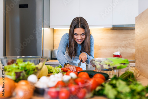 Delightful and happy young woman stands and cooks in kitchen. She cuts lettuce on wood board. Girl has many ingrediends on table.