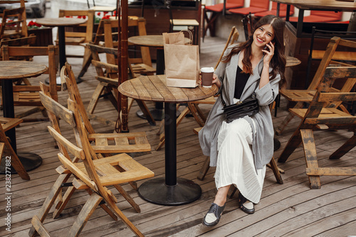 Stylish hipster girl with beautiful hair and smile talking on phone, with coffee cup and bag on wooden table on terrace in city street. Gorgeous happy young woman enjoying time and smiling