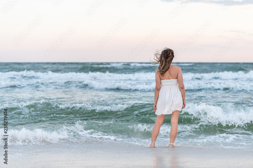 Young woman back standing in white dress on beach sunset in Florida panhandle with wind, ocean waves, tan skin Young woman back standing in white dress on beach sunset in Florida panhandle with wind, ocean waves, tan skin
