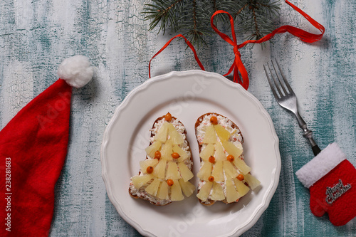 New-year sandwiches with cheese, pineapples and crab stiks in the form of a Christmas tree, are located on a white plate. Sandwiches are decorated with buckthorn berries photo