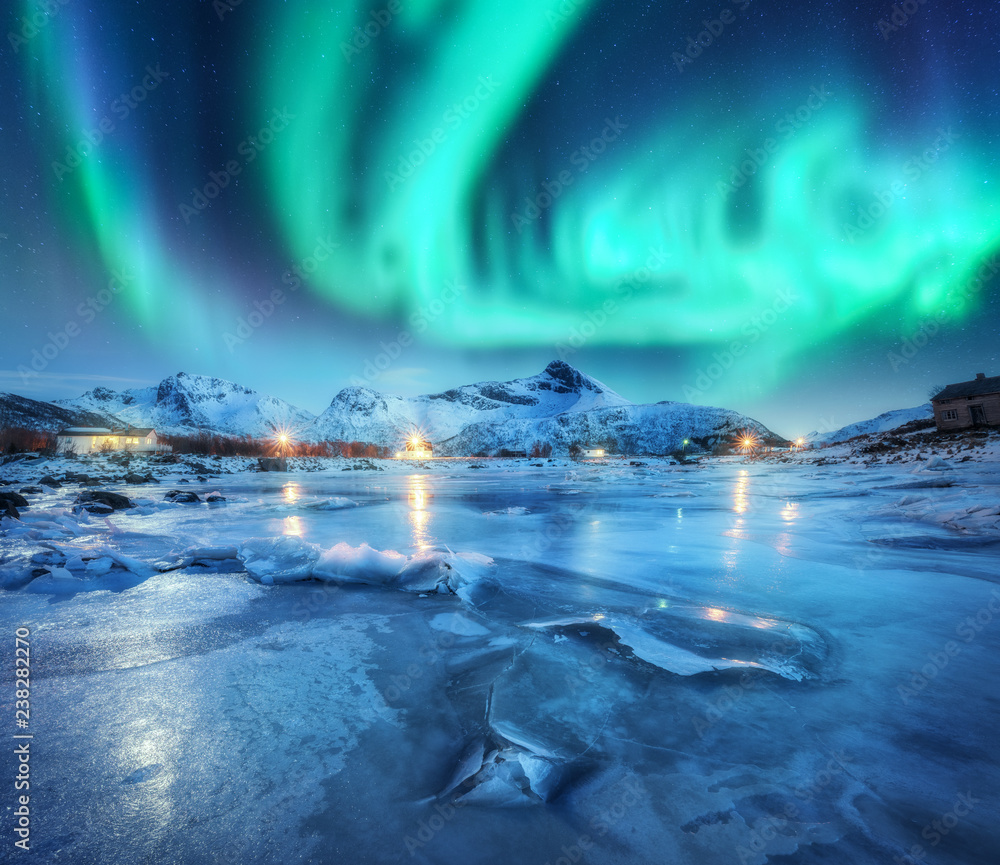 Northern lights above snowy mountains, frozen sea coast and houses in Lofoten islands, Norway. Aurora borealis and small village. Winter landscape with polar lights, rorbu, ice. Aurora over rocks