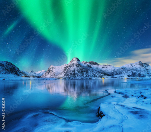 Aurora borealis over snowy mountains, frozen sea coast and reflection in water in Lofoten islands, Norway. Northern lights. Winter landscape with polar lights, ice in water. Starry sky with aurora