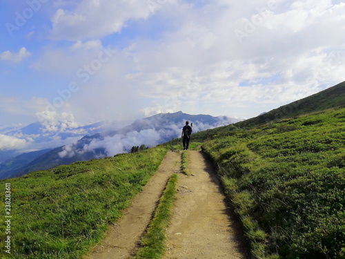 Escursionista cammina su sentiero di montagna con sullo sfondo montagne e cielo nuvoloso © Sandro