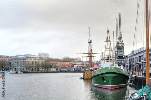 A view of the Bristol wharf on Avon river from Spike Island, with two green sailing boats and three cranes in a cloudy winter day, in United Kingdom