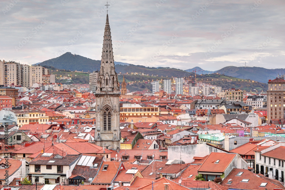 A landscape of Bilbao mountains and city centre with clouds, mountains, houses roofs and the cathedral belltower at sunset, in Vizcaya, Spain