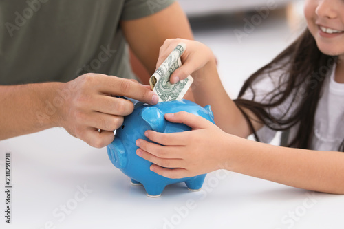 Little girl with her father putting money into piggy bank at table, closeup