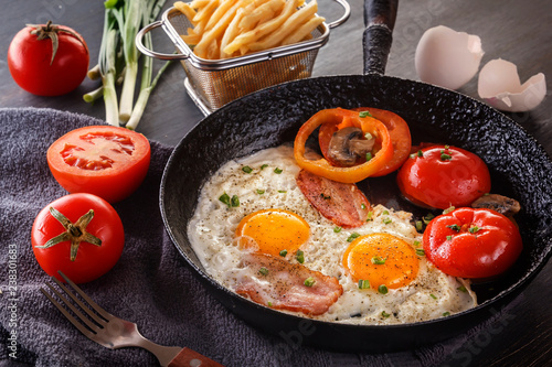 Fried eggs with bacon and tomatoes on an old cast-iron pan with french fries on a gray table. Close-up