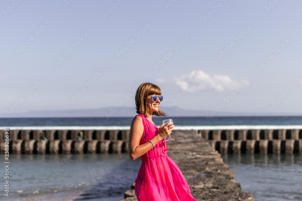Young woman in a long red dress and with a glass of wine posing on the sea background. Young woman in a long red dress and with a glass of wine posing on the sea background.