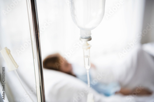 Selective focus at IV tube stand  with blurred sick girl lying on the hospital bed.