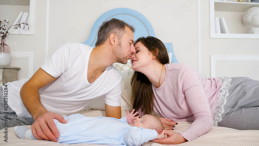 Attractive man and woman kissing while lying on soft bed near sweet sleeping baby in stylish bedroom. Attractive man and woman kissing while lying on soft bed near sweet sleeping baby in stylish bedroom.