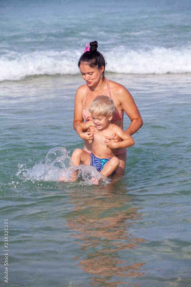 beautiful mom brunette is playing with her son blonde against the sea on the beach