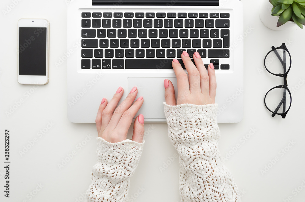 Φωτογραφία Top view of woman's hands typing on laptop keypad placed on white office desktop with blank smartphone, glasses and decorative plant