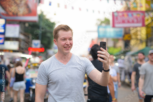 Young happy tourist man taking selfie against view of busy street in Khao San in Bangkok